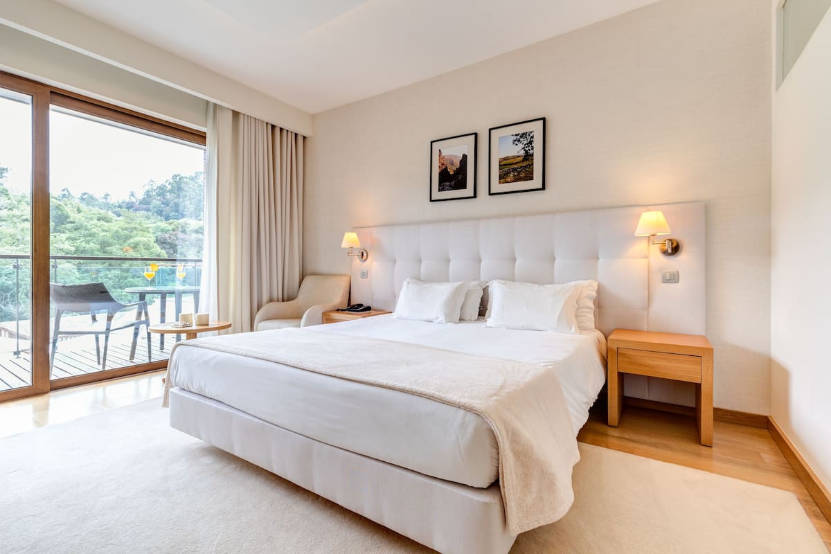 A large bed with a headboard, two bedside tables with lamps, a chair, and a balcony with a table and chairs.