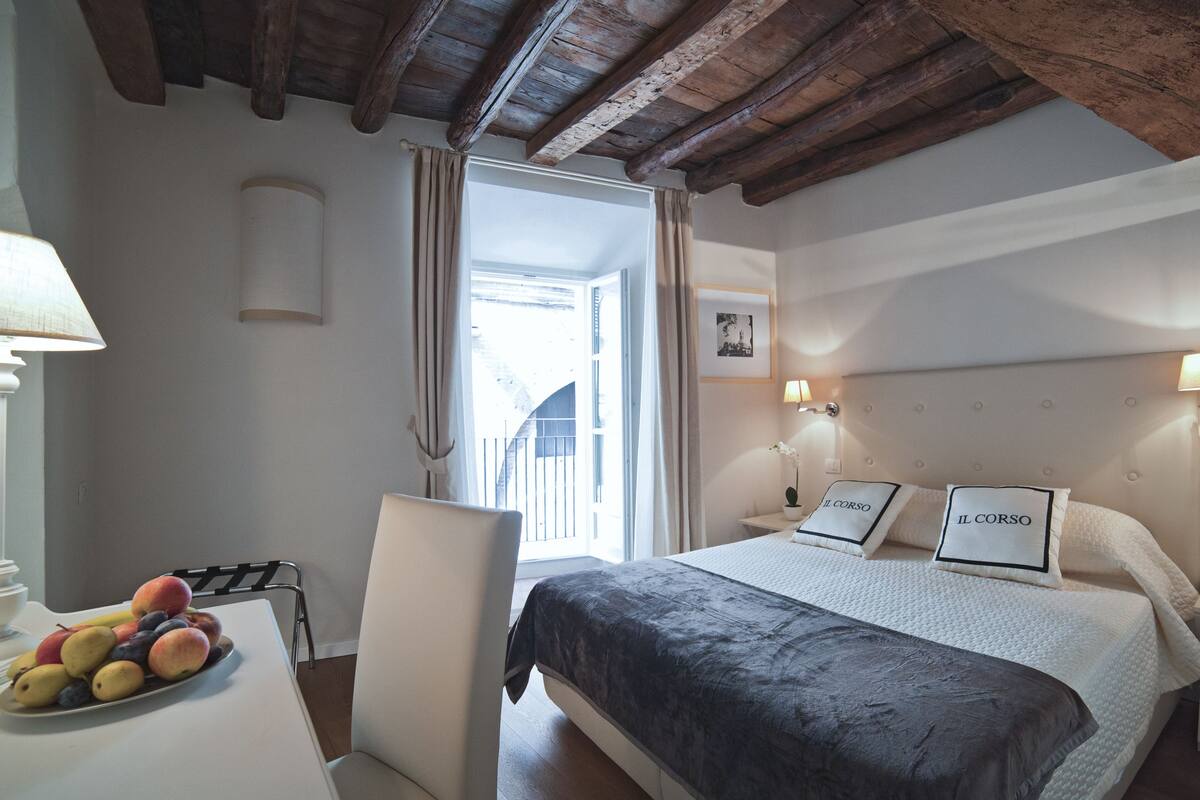 Habitación de hotel con cama, escritorio con frutero, una silla, ventana con cortinas y techo de madera.