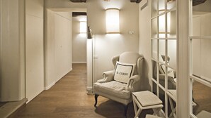 A hallway with a wooden ceiling, a white armchair with a pillow labeled "IL CORSO", a floor lamp, and a small table with a book. - B&B Il Corso (Siena)