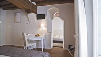 A bedroom with a bed, a desk with a lamp, a chair, and a small table with fruit. There is a TV on the wall and a window with curtains.