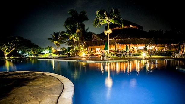 Vista nocturna de una piscina con un deck de piedra, palmeras y un edificio con techo de paja al fondo.