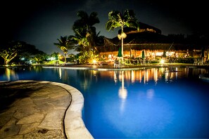 Vista nocturna de una piscina con un deck de piedra, palmeras y un edificio con techo de paja al fondo.