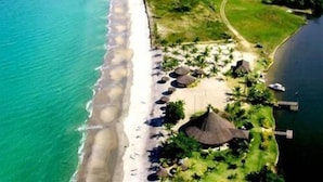 Vista aérea de un complejo turístico en la playa, con una extensa playa de arena, algunas estructuras con techo de paja y una pequeña masa de agua.