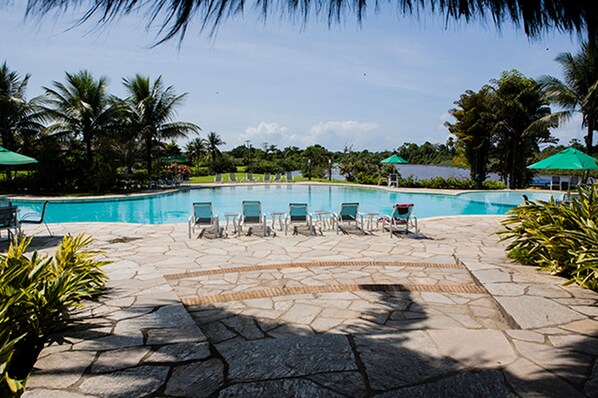 Un área de piscina con sillas de descanso y sombrillas, rodeada de palmeras y un cielo despejado.