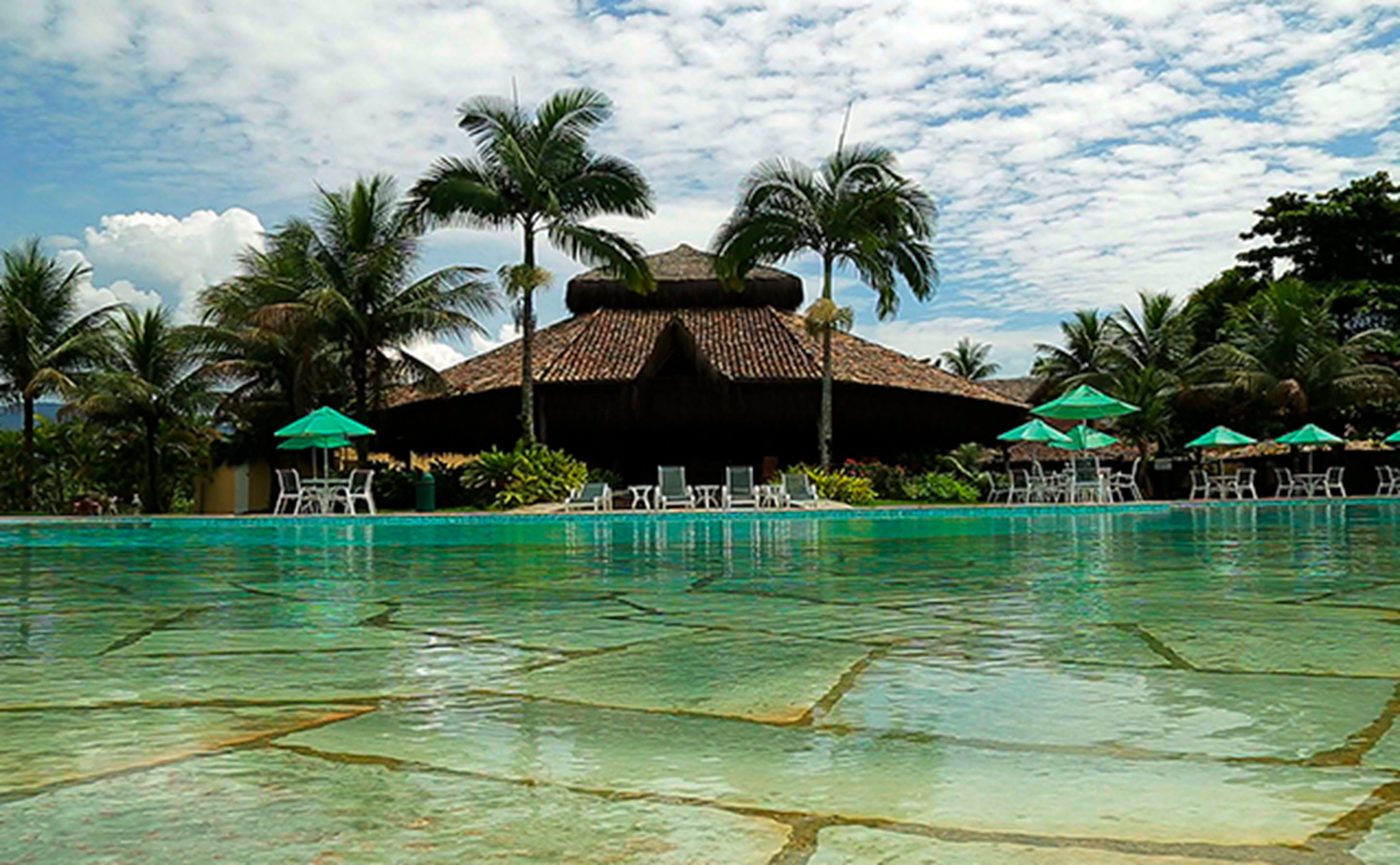 Un espace piscine agrémenté de chaises longues et de parasols, entouré de palmiers et bordé d’un bâtiment à toit de chaume.
