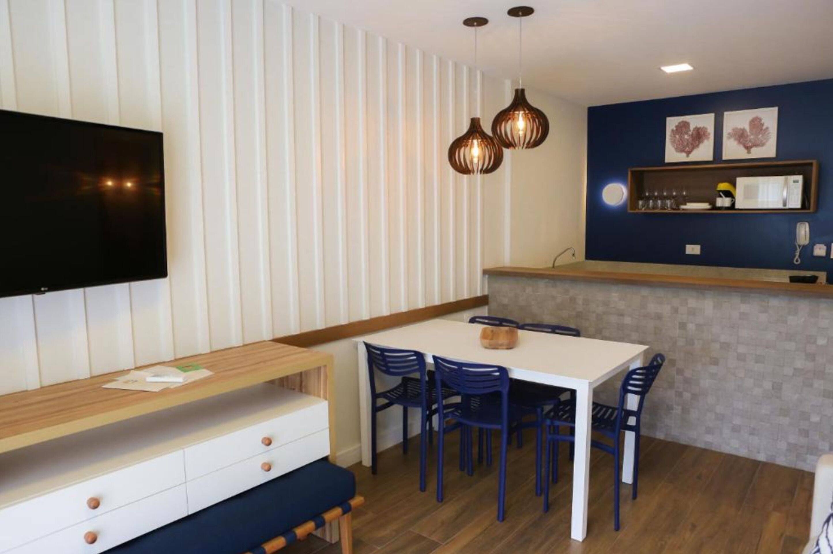 Una cocina moderna con encimera blanca, sillas azules y un banco de madera. Hay un televisor montado en la pared y lámparas colgantes sobre la encimera.