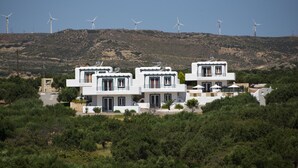 A modern white building with multiple levels and balconies, surrounded by dense greenery and wind turbines in the background. - Cavo Plako Villas (Sitia)