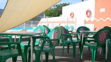 Outdoor seating with green plastic chairs and tables, a shaded area, and a building with a lifebuoy on the wall.
