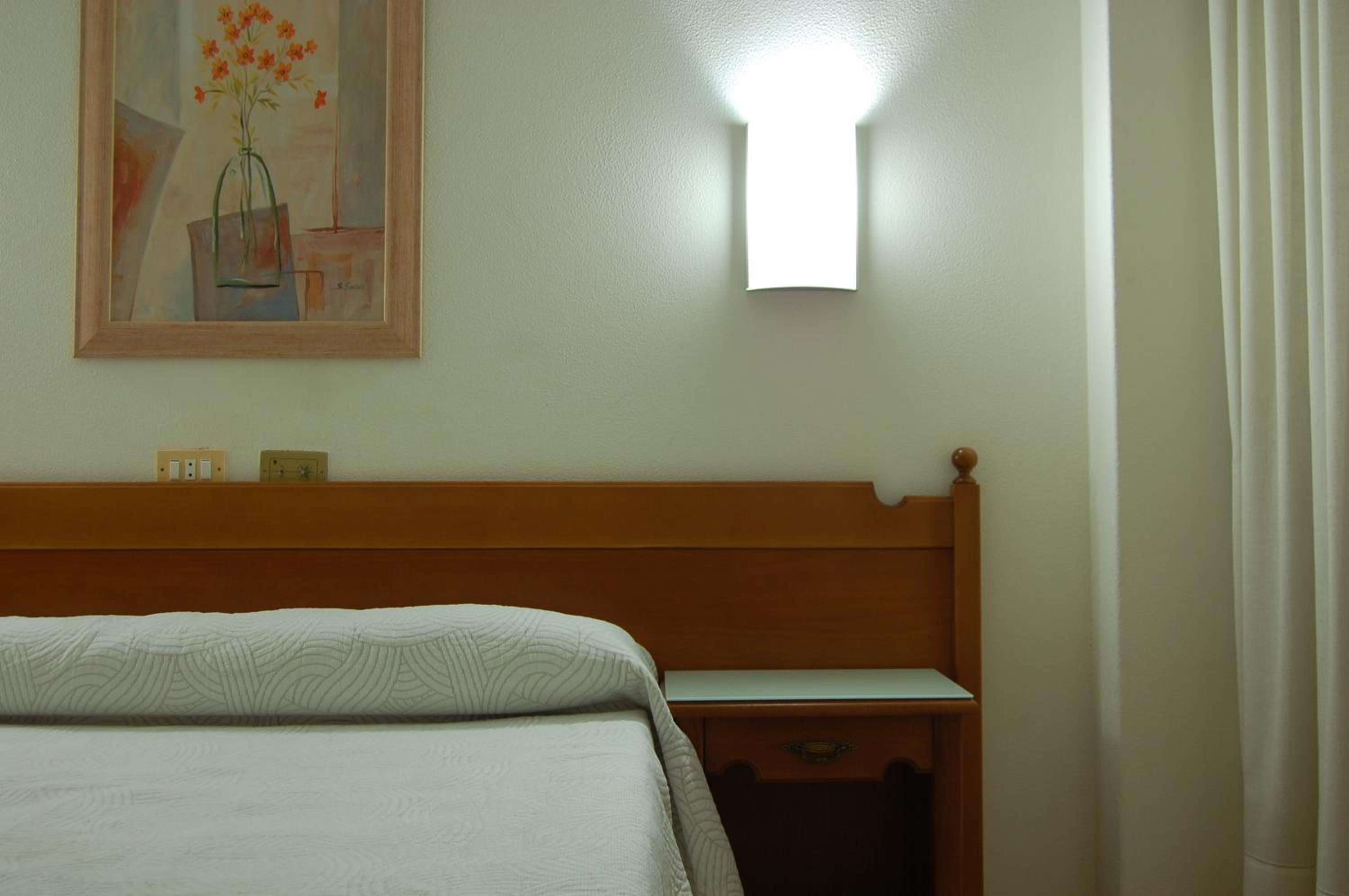 Una habitación de hotel con una cama, una mesita de noche, una lámpara de pared y un cuadro sobre la cama.