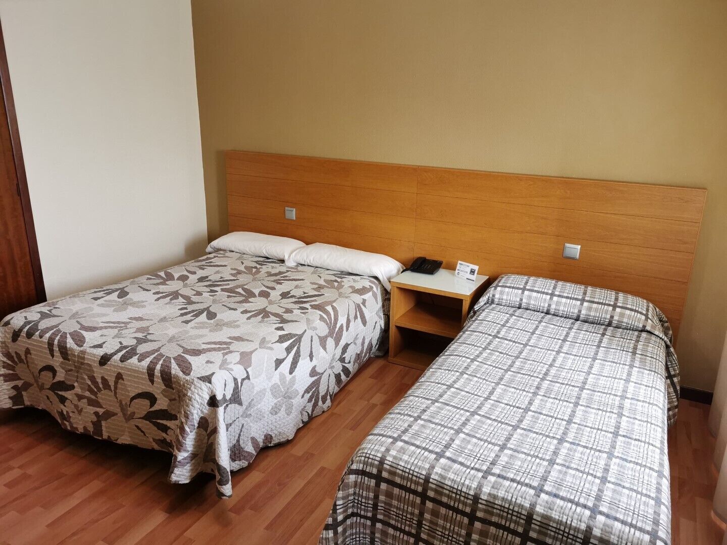 A hotel room with two beds, a wooden headboard, a bedside table with a phone and a sign, and a wooden floor.
