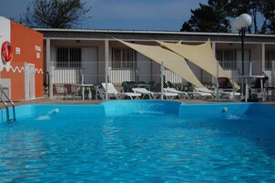 Una piscina con sillones de descanso y una zona sombreada, rodeada por una valla y un edificio con un flotador en la pared.