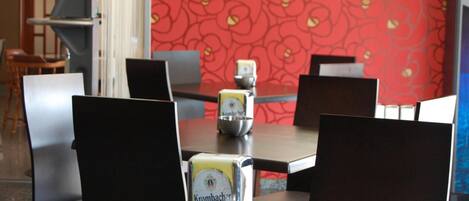 A restaurant with a red floral wall, black chairs, and tables with Krombacher beer glasses.