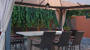 An outdoor patio with a stone table and wicker chairs, surrounded by a pergola with climbing vines.