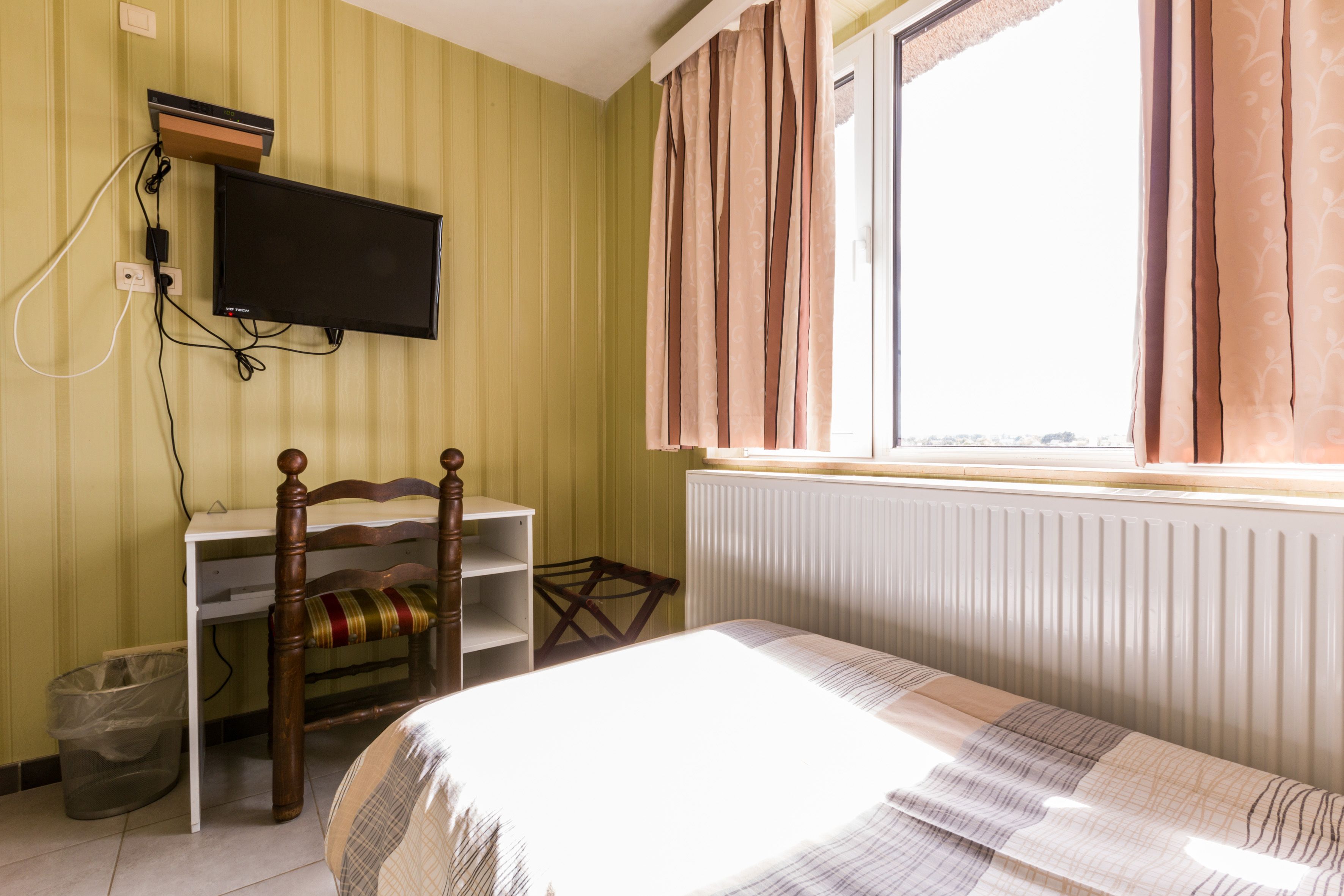 A room with a bed, a desk with a chair, a television mounted on the wall, and a window with curtains.