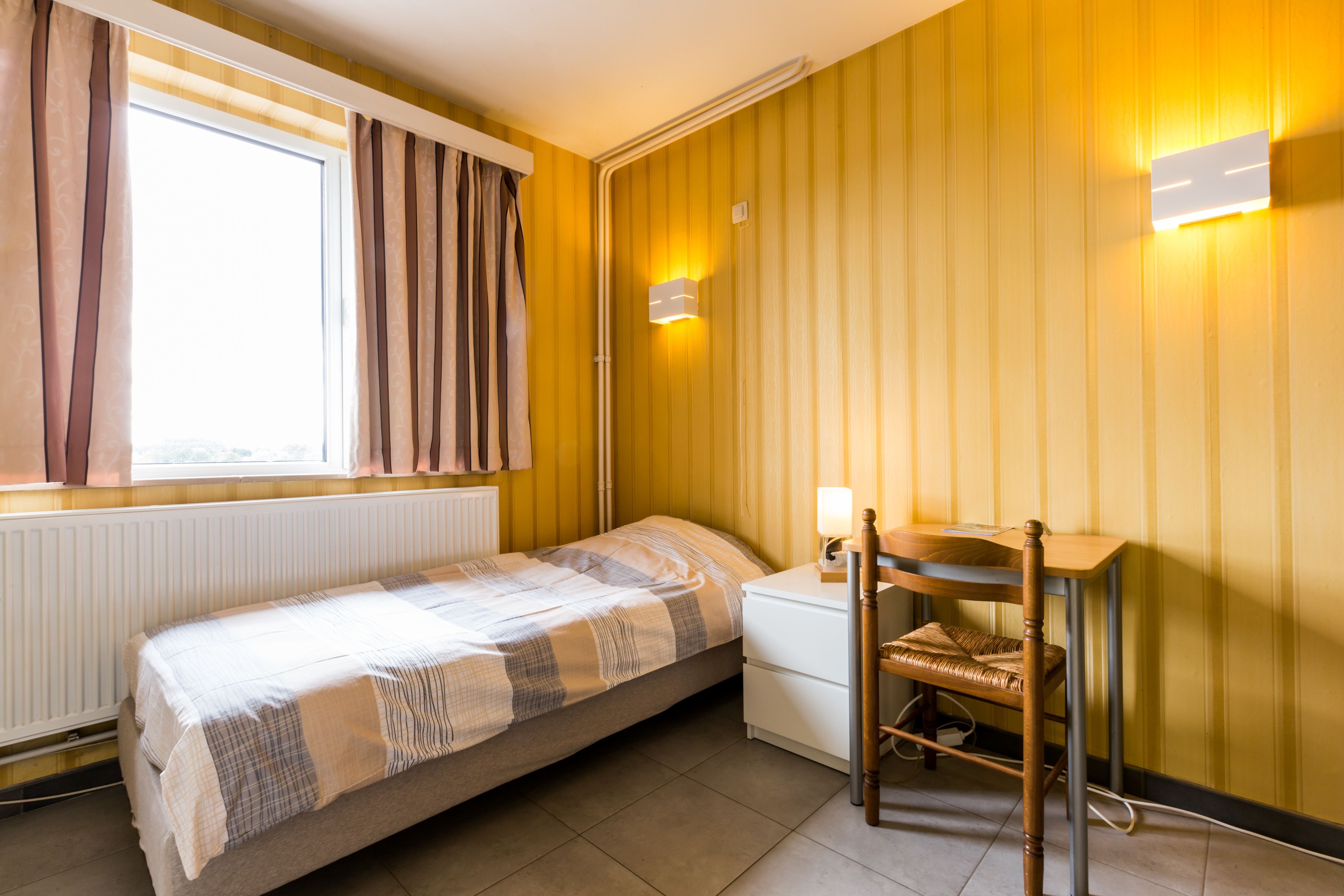 A small, single-bed room with a window, a bedside table with a lamp, a chair, and a wall-mounted light fixture.