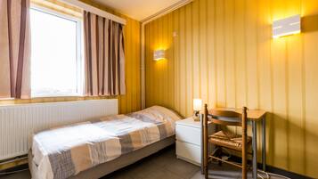 A small, single-bed room with a window, a bedside table with a lamp, a chair, and a wall-mounted light fixture.