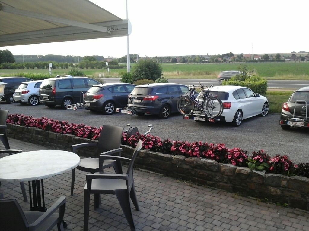 Uma área de estacionamento com vários carros e uma bicicleta. Há mesas e cadeiras ao ar livre.
