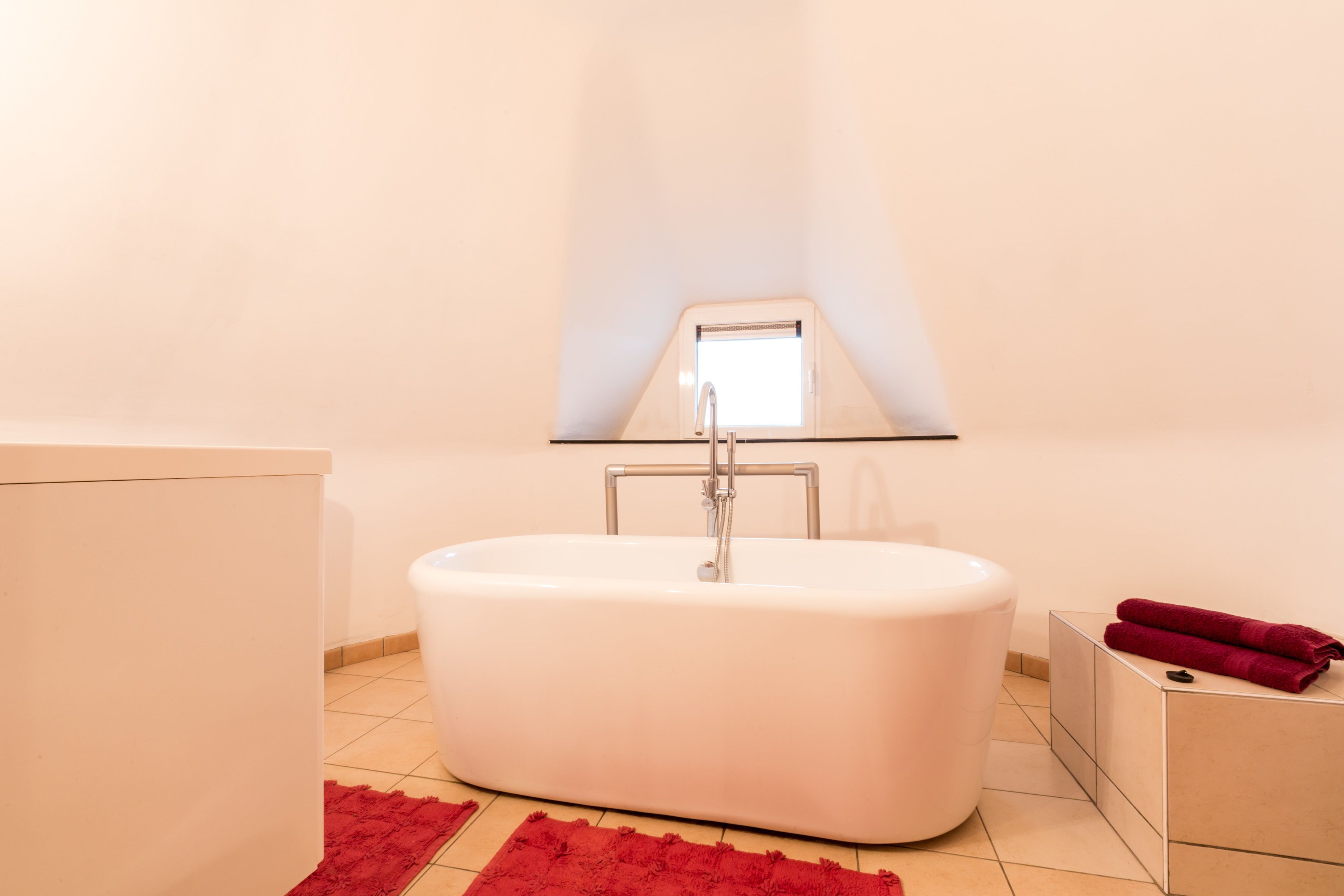 A modern bathroom with a freestanding bathtub, a small window, and a red rug on the floor.