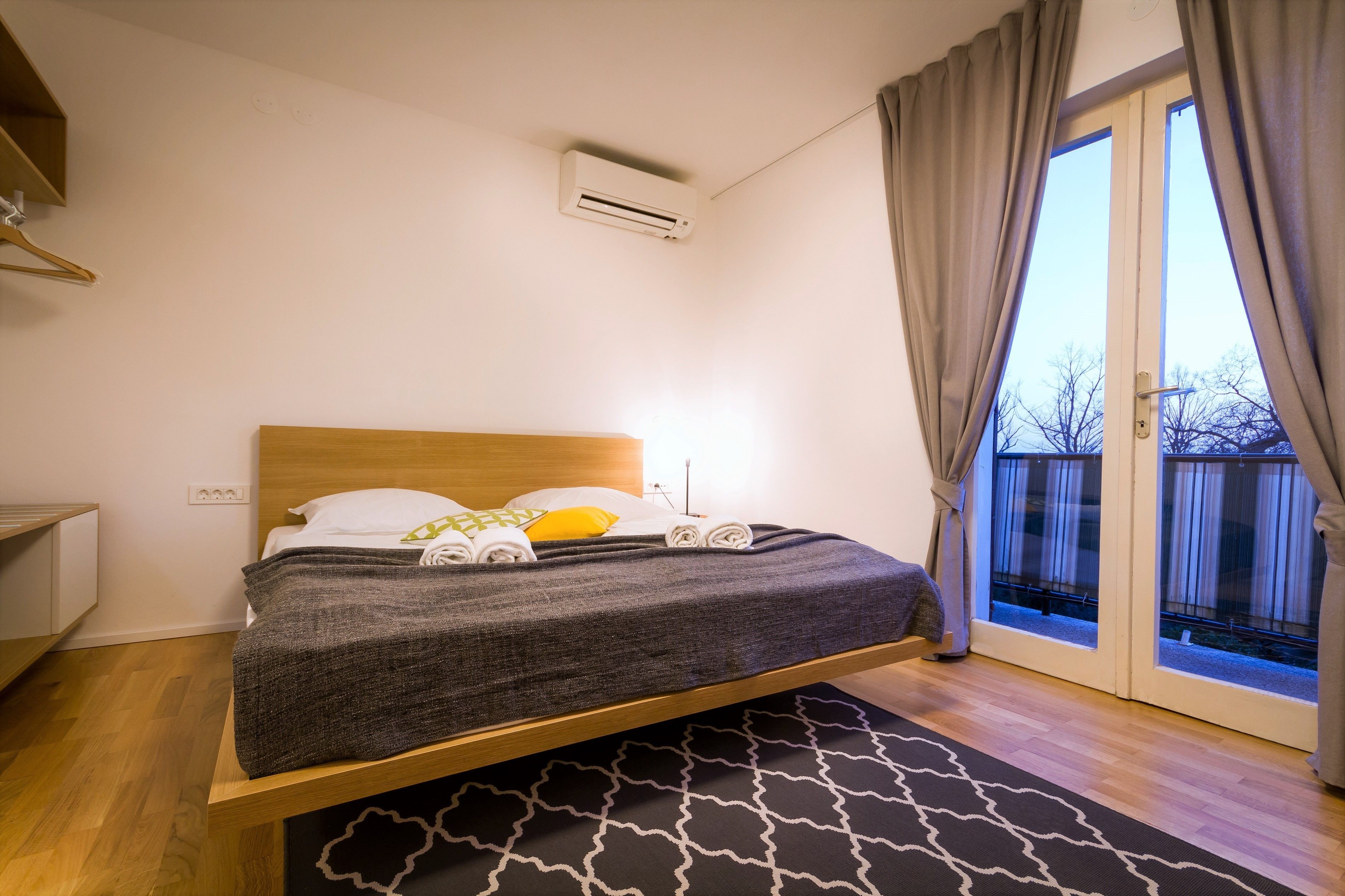 A bedroom with a wooden bed, a small bedside table, a wall-mounted air conditioner, and a view of a balcony through a glass door.