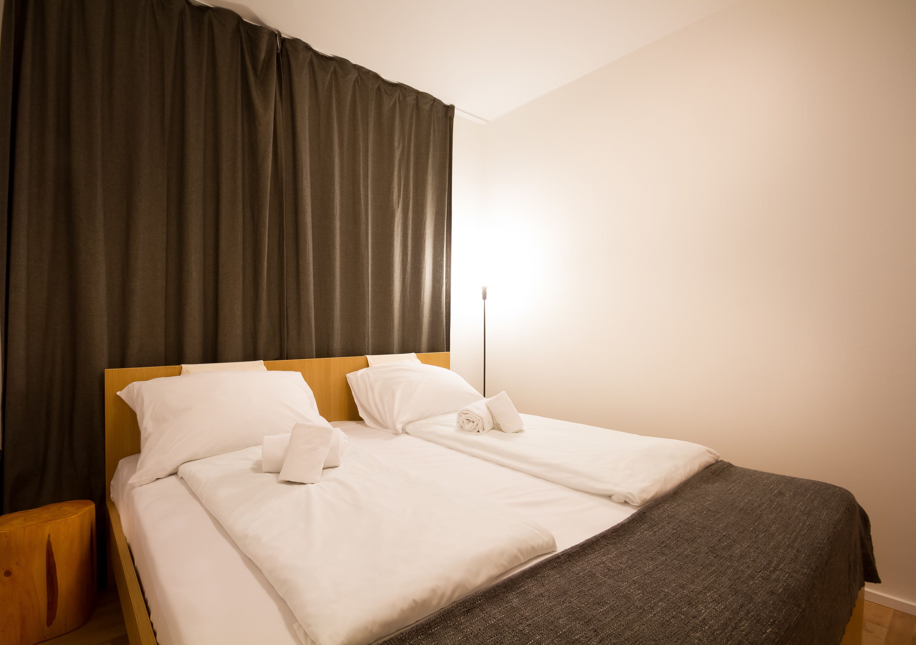 Una cama bien tendida con sábanas blancas y una manta marrón oscuro, una mesita de noche de madera y un ventanal con cortinas.