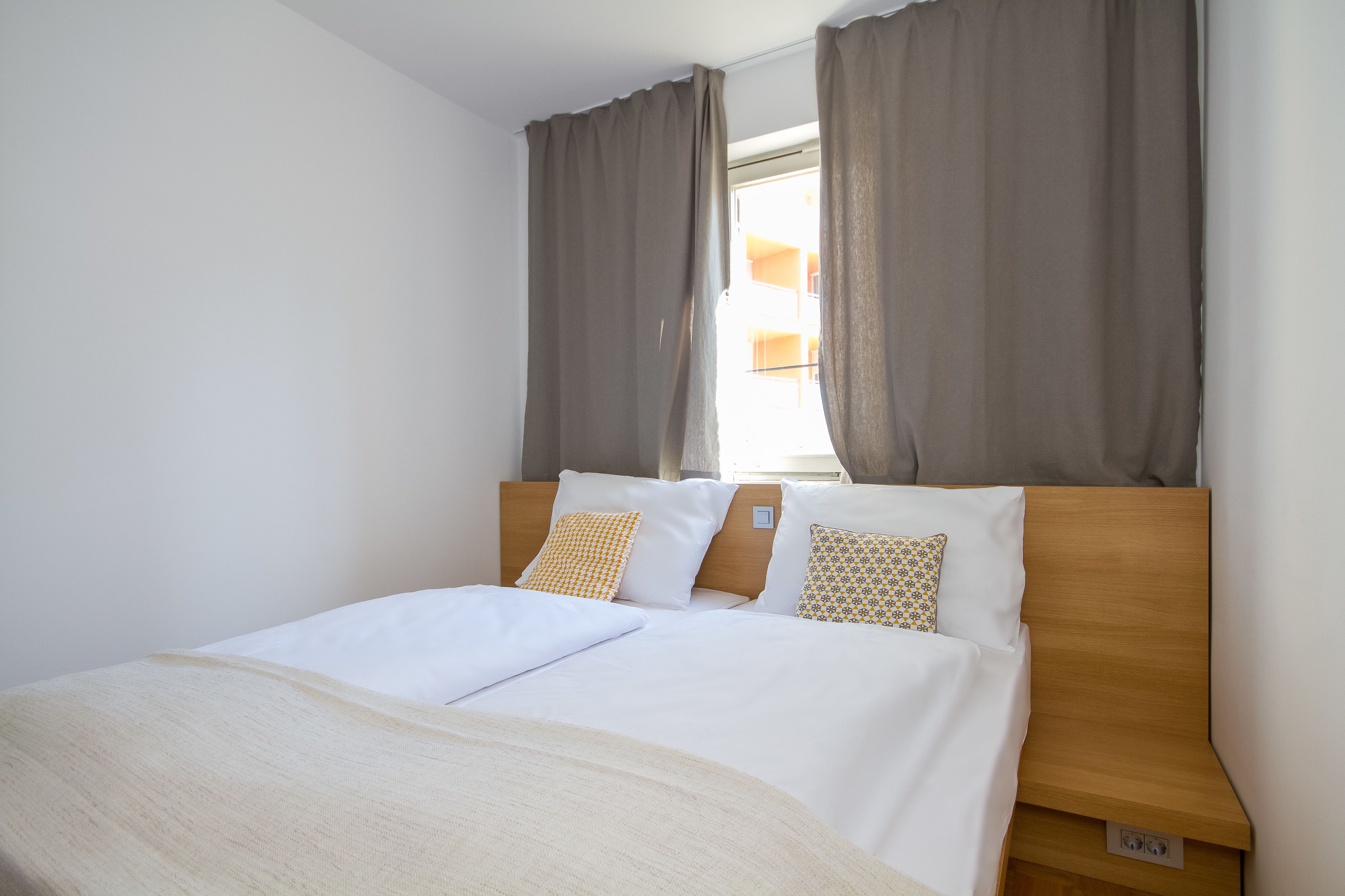 A neatly made bed with white linens and two patterned pillows, a wooden headboard, and a window with curtains.