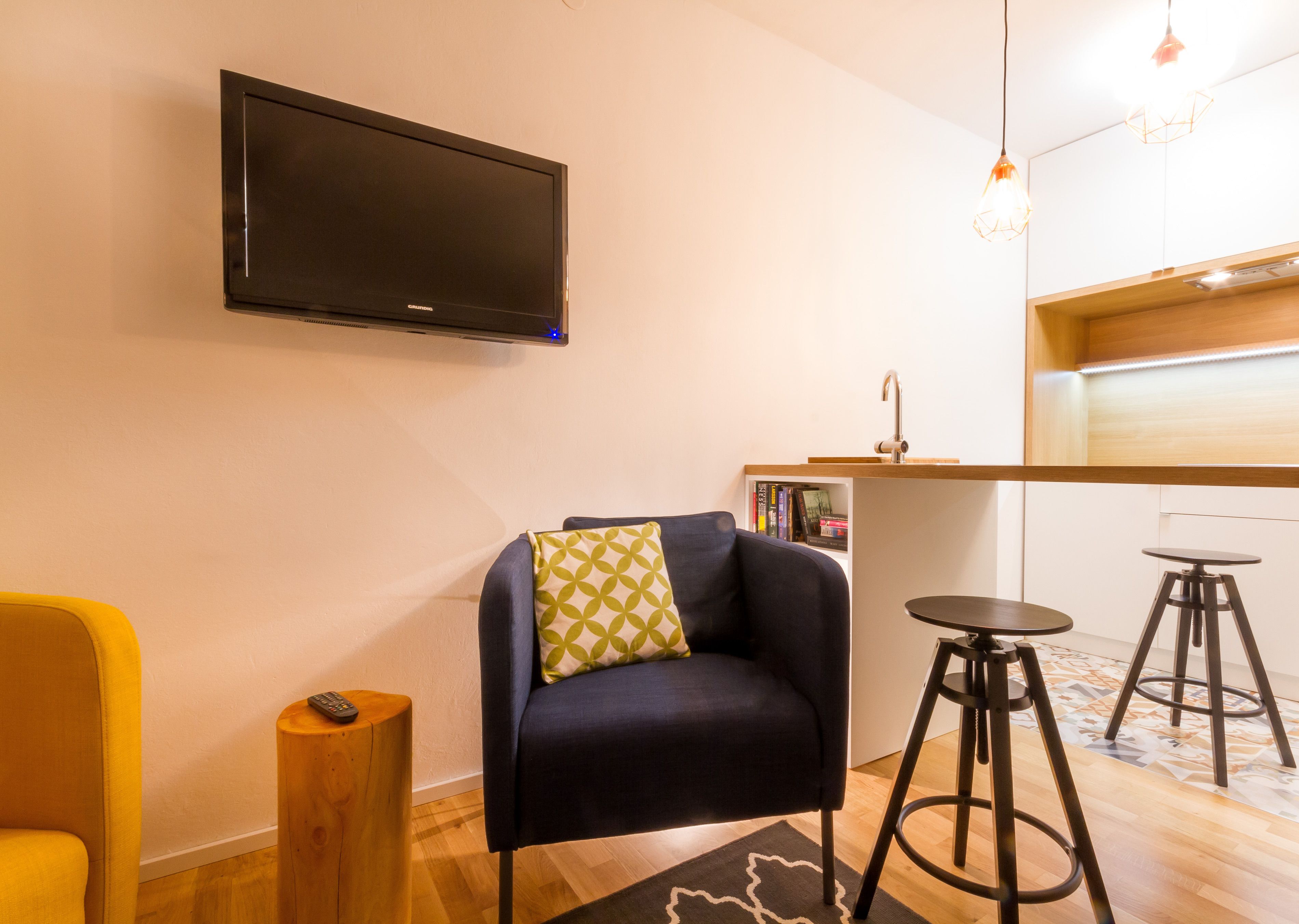 A modern living room with a flat-screen TV mounted on the wall, a dark blue armchair with a patterned pillow, a yellow armchair, a wooden stool, and two bar stools.