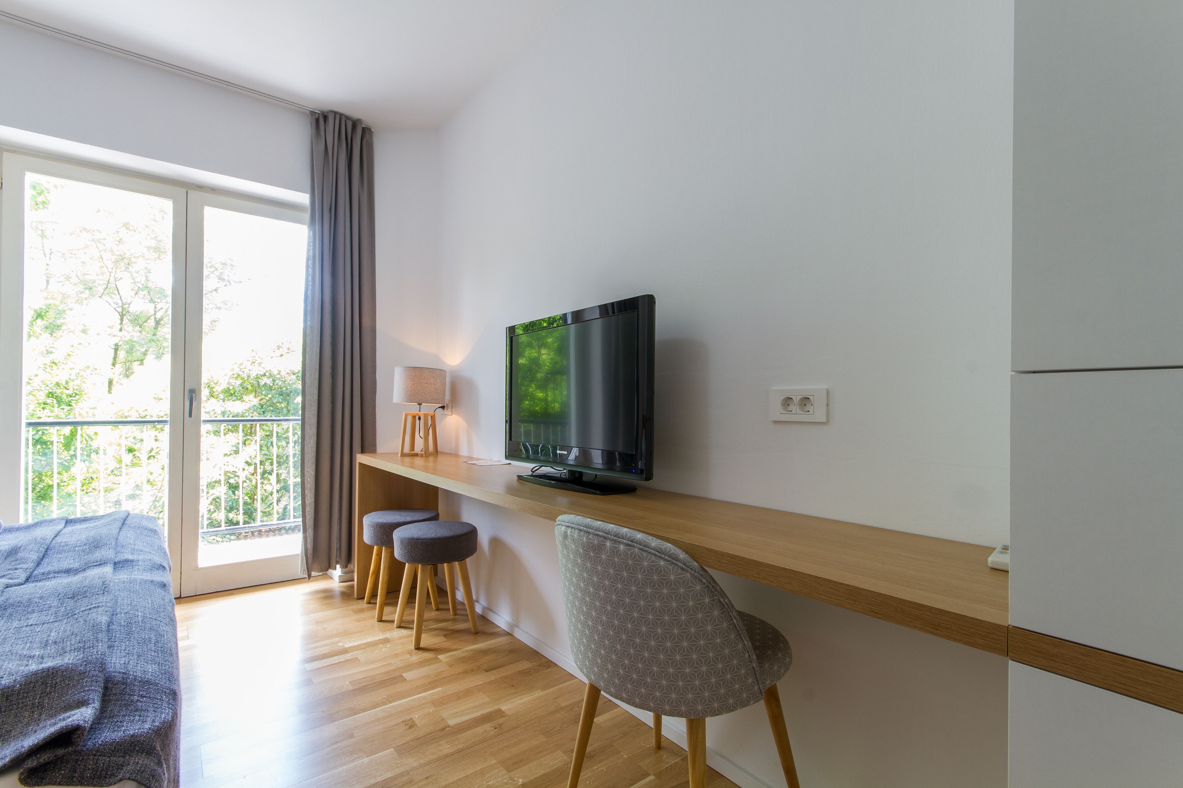 A modern room with a TV on a wooden shelf, a bed, a desk with a chair, and a balcony with curtains.