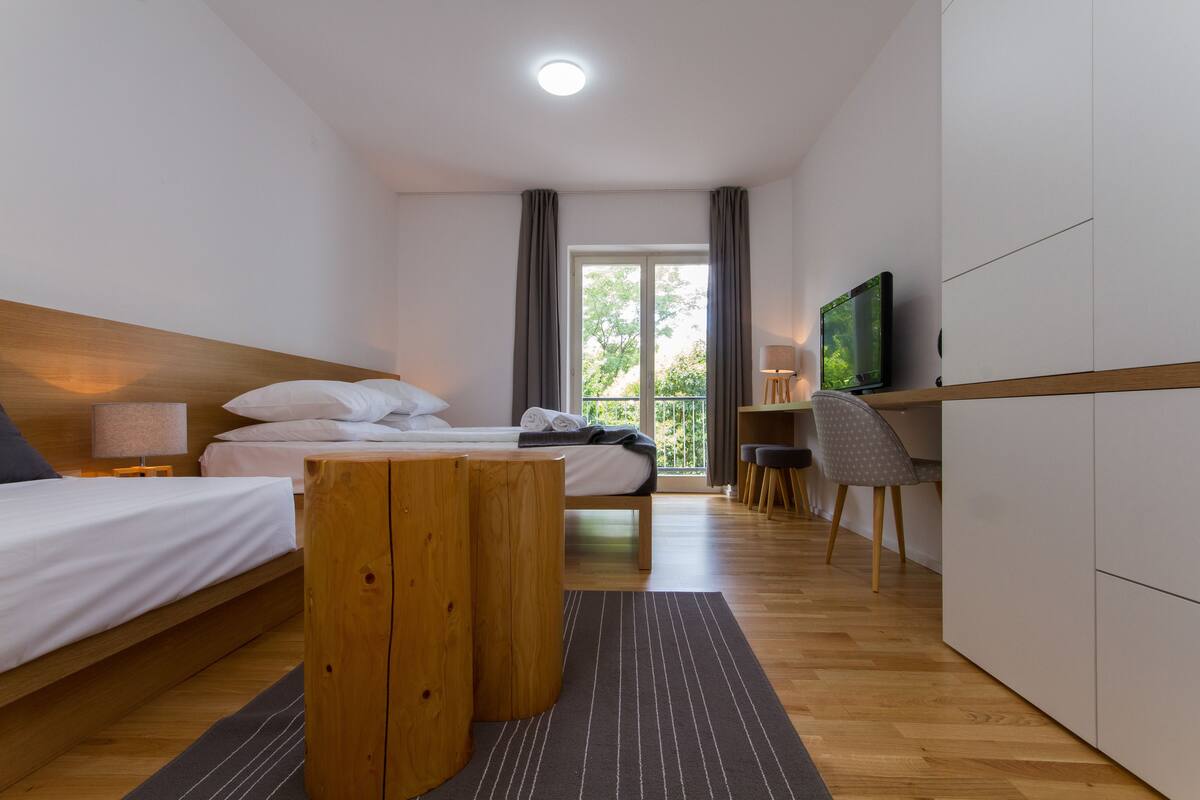 A modern hotel room with a wooden bed, a desk, a chair, a TV, and a balcony with a view.