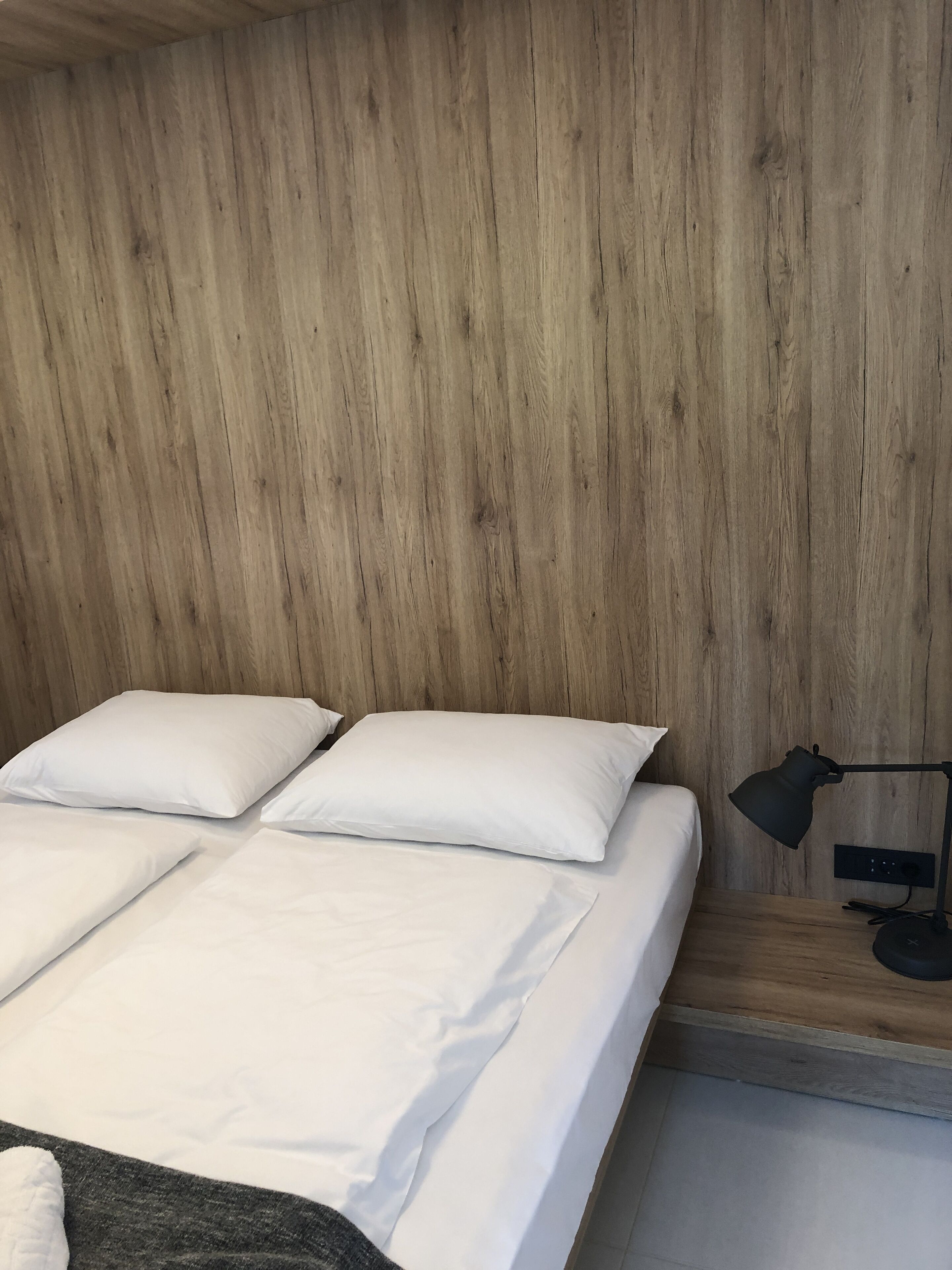 Una cama con ropa de cama y almohadas blancas, una mesita de noche con una lámpara y una pared de madera.
