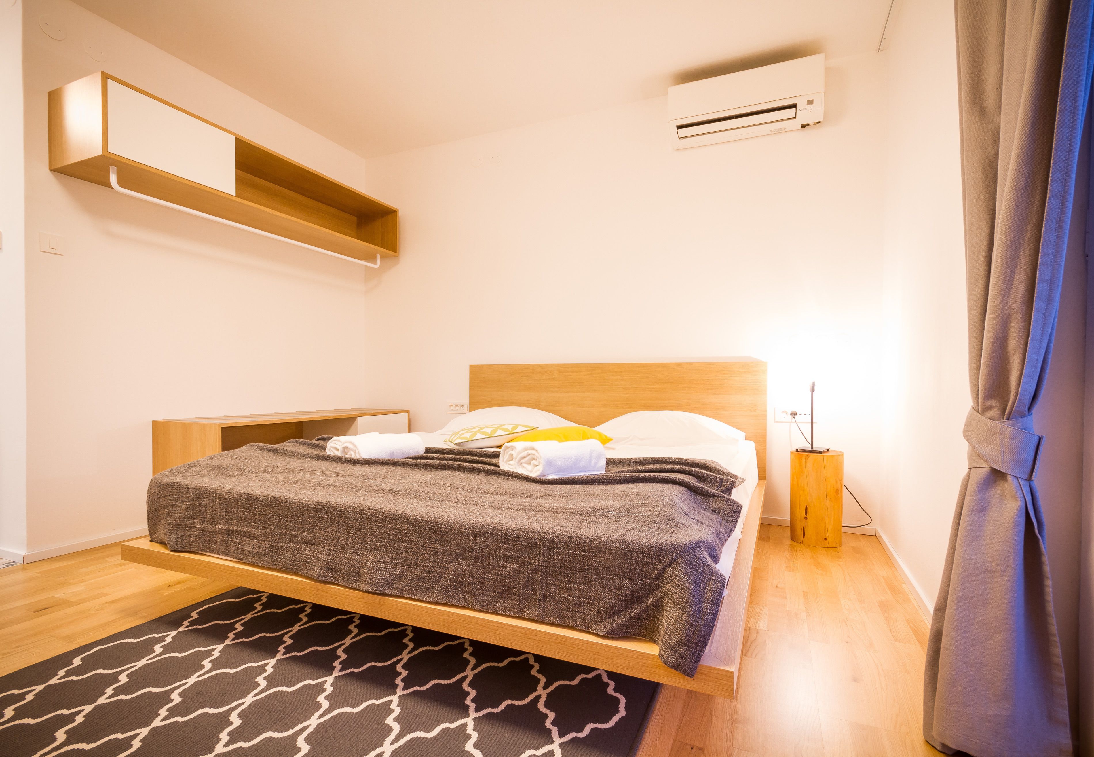 Een netjes opgemaakt bed met een grijze deken, een houten hoofdbord en een nachtkastje met een lamp.