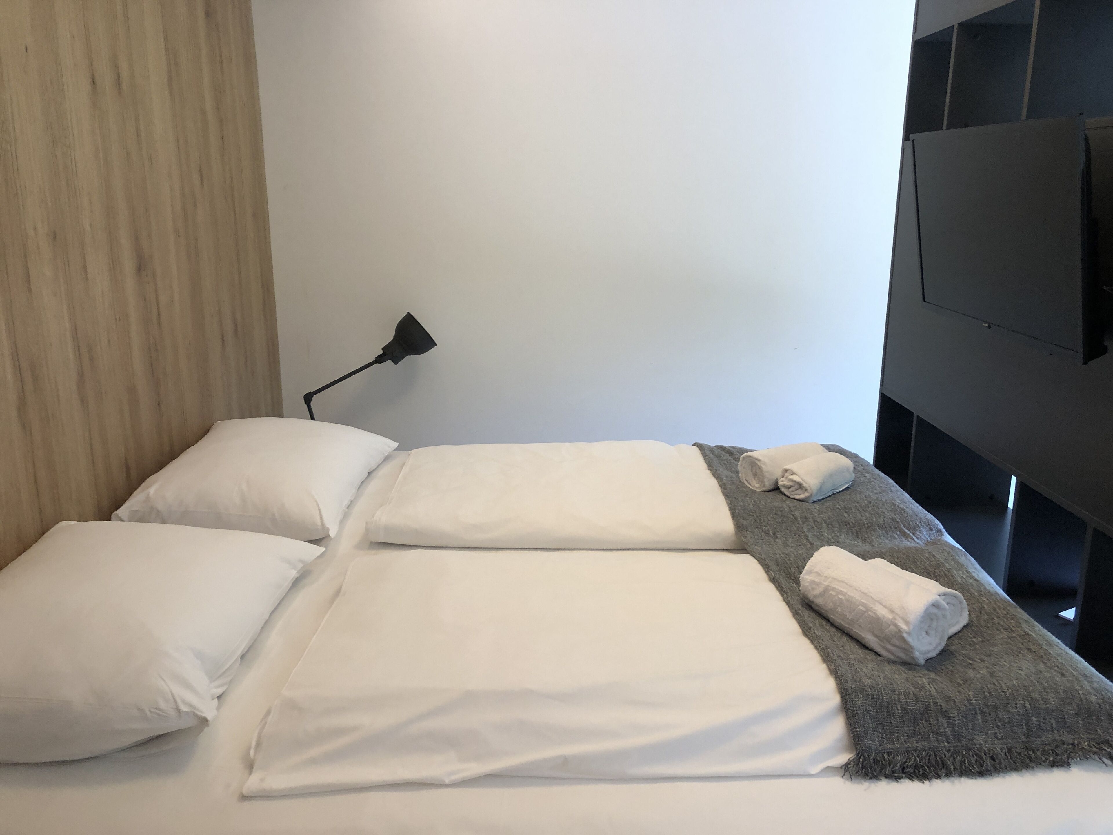 A neatly made bed with white linens, a grey throw blanket, and rolled towels on a dark headboard. A black desk lamp is positioned on the wall to the left of the bed.