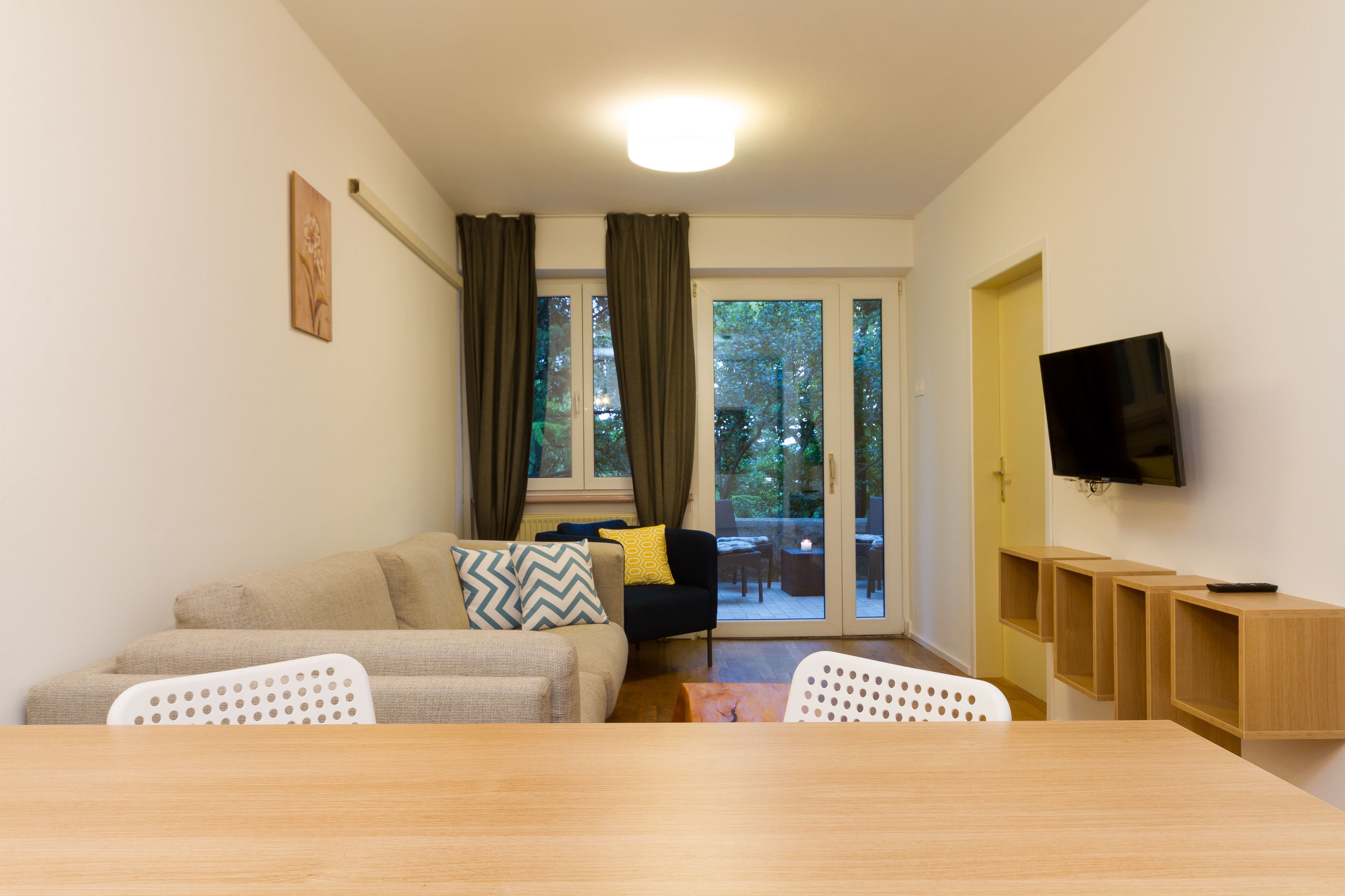 Ein Wohnzimmer mit einer Couch, Sesseln, einem Tisch, einem Fernseher und einer Glastür, die zu einem Außenbereich führt.