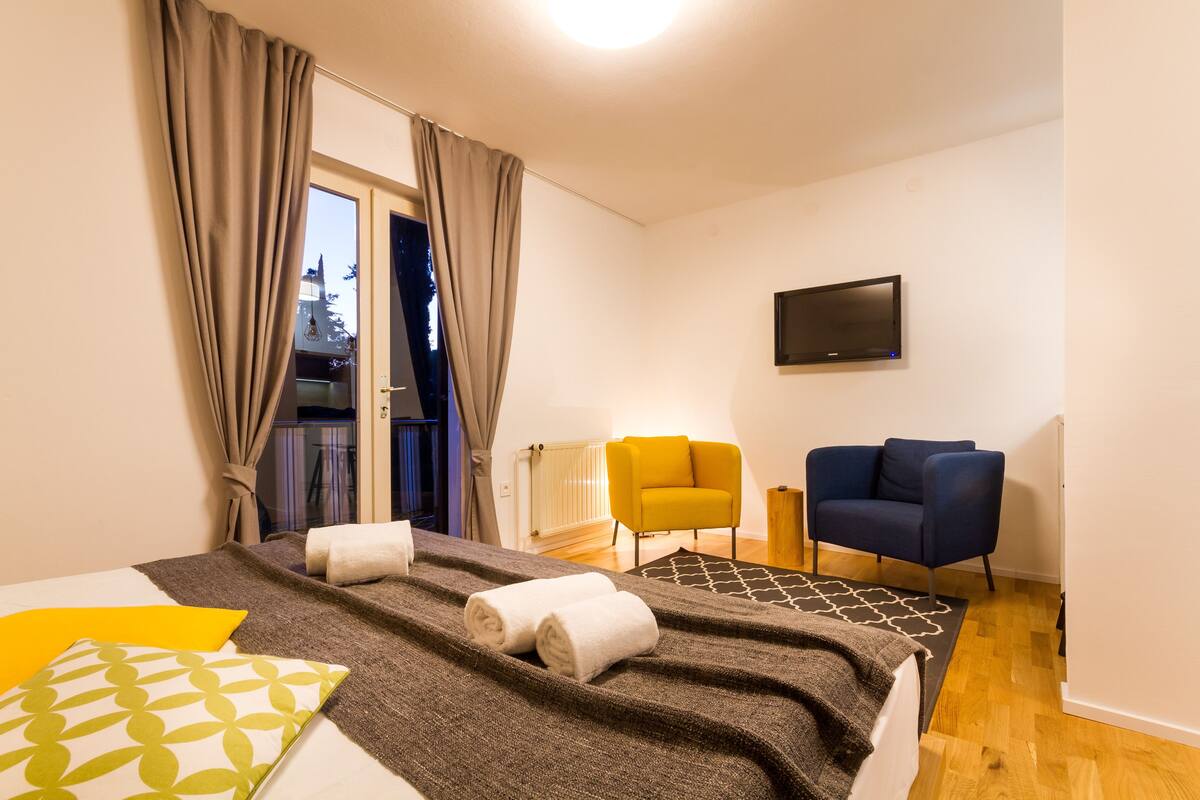 A hotel room with a bed, a TV, a yellow armchair, a blue armchair, and a view of the outside.