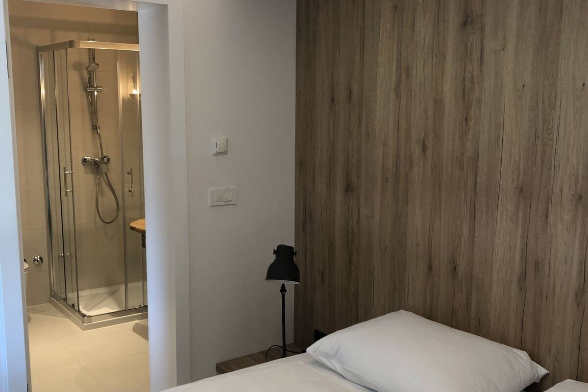 A hotel room with a wooden headboard, a bed with white linens, a glass shower enclosure, and a bedside lamp.