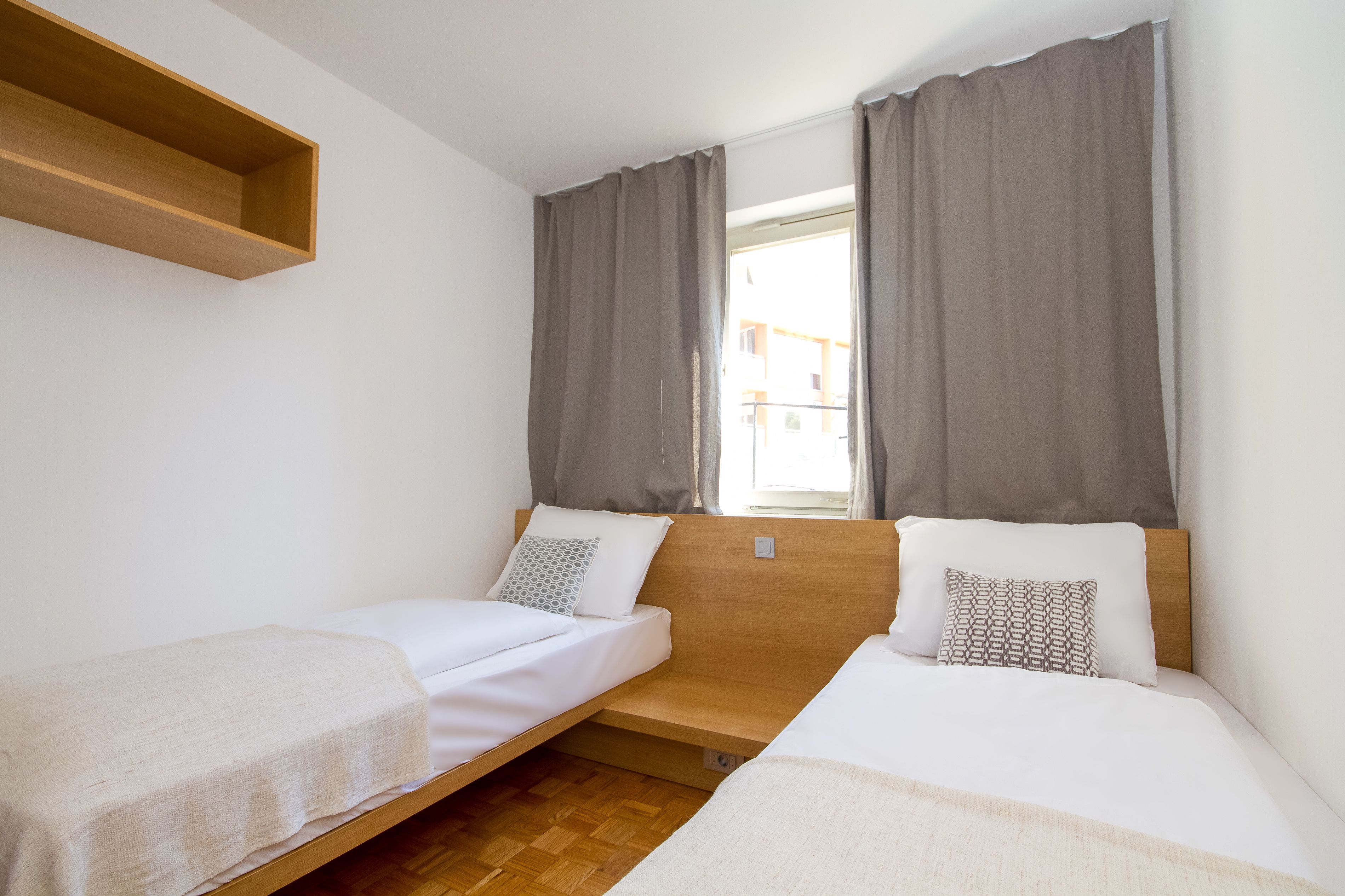 A room with two single beds, a wooden shelf, and a window with curtains.