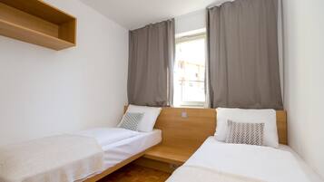 A room with two single beds, a wooden shelf, and a window with curtains.