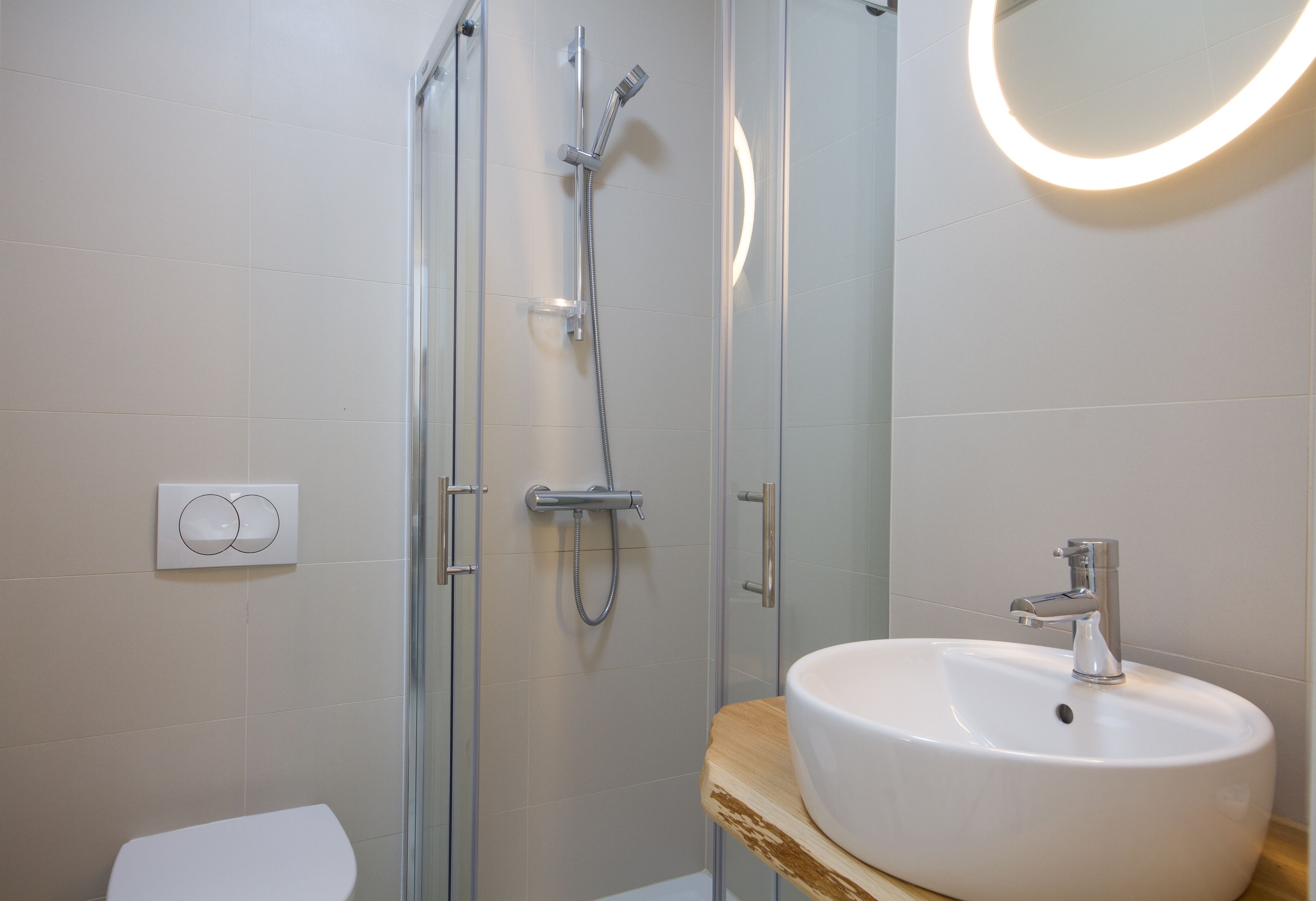 A modern bathroom with a glass-enclosed shower, a white sink with a wooden countertop, and a round mirror with a light.