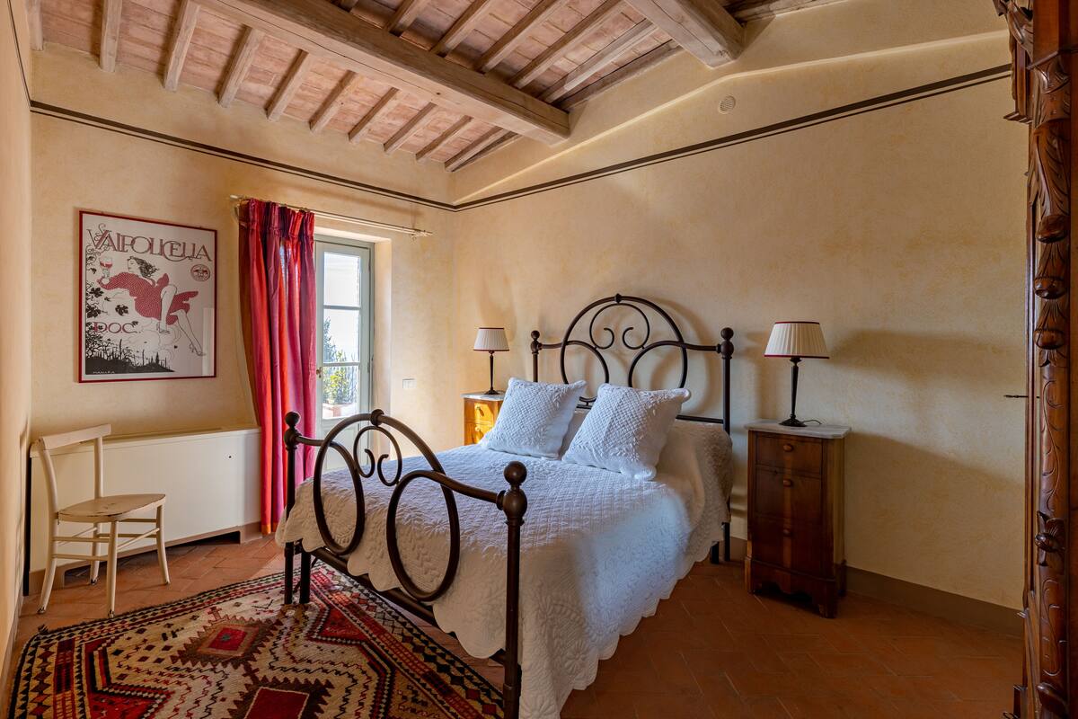 A bedroom with a bed, two bedside tables, a chair, a window with curtains, and a wall-mounted picture.