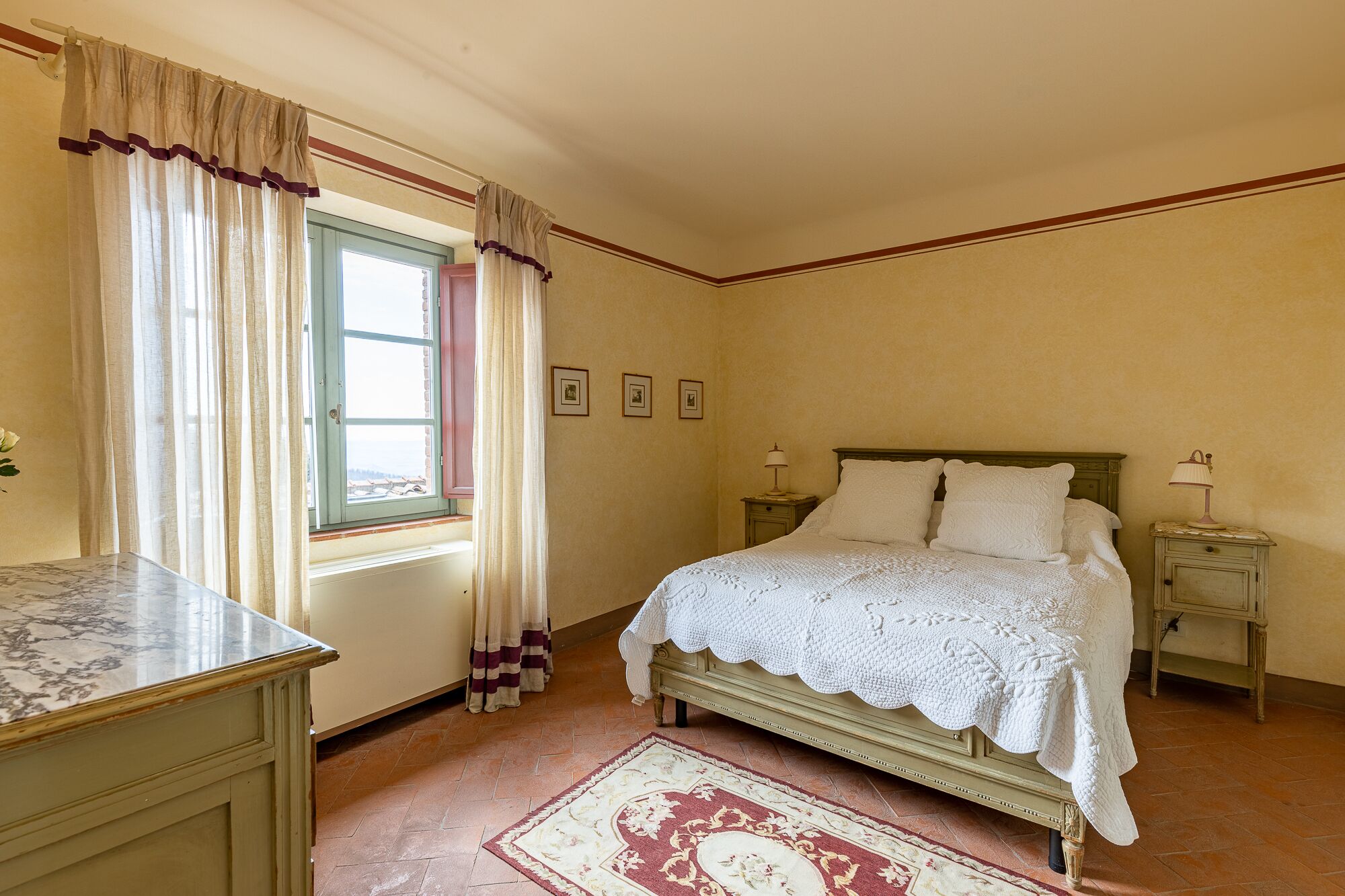 A bedroom with a bed, bedside tables, a nightstand, a window with curtains, and a marble table.