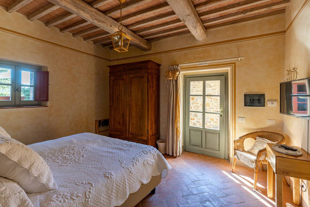 A bedroom with a bed, wooden wardrobe, a door with a stone window, a wicker chair, and a small table with a remote control.