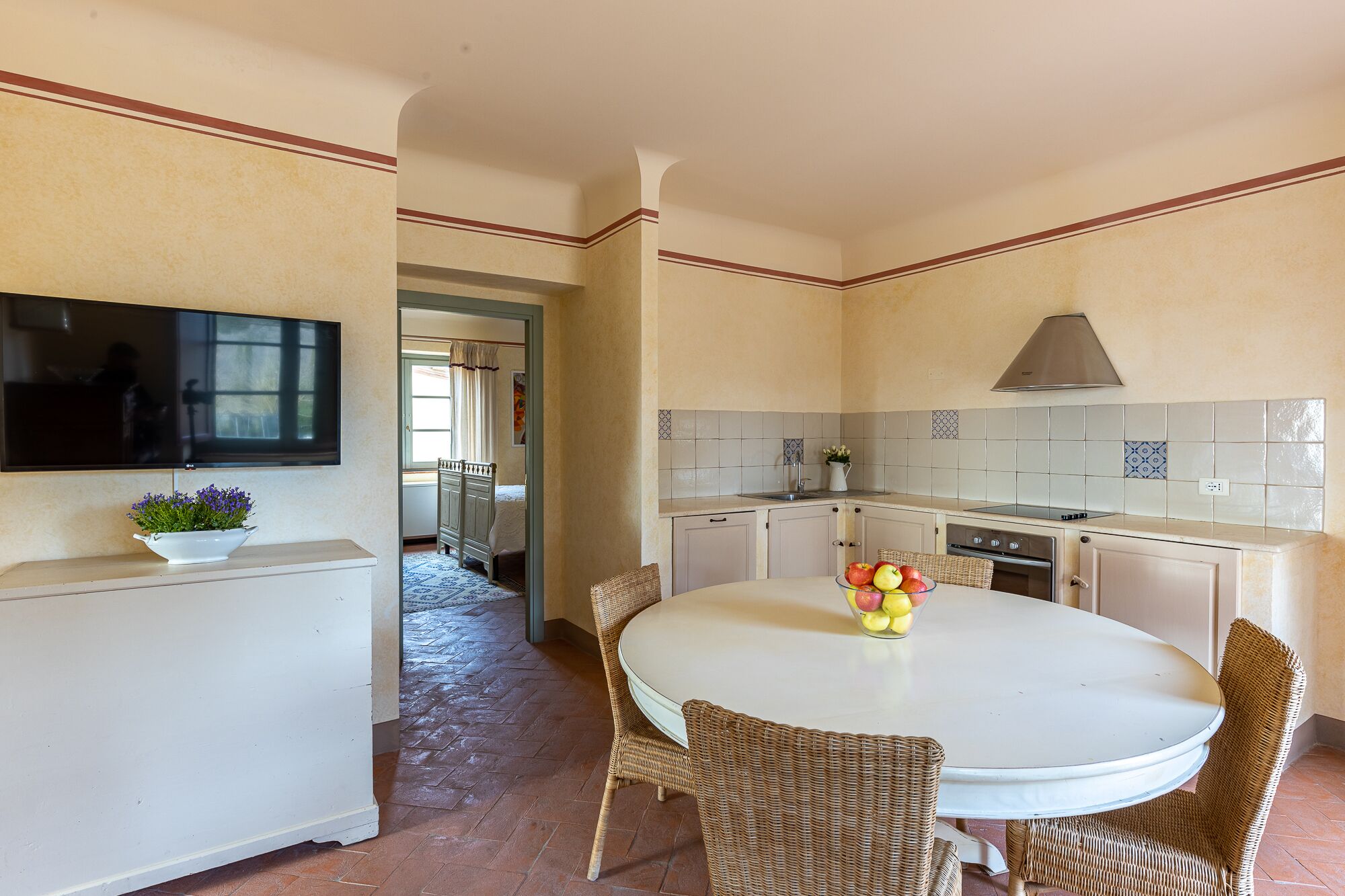 A compact kitchen with a dining area, a TV mounted on the wall, and a small table with fruit.