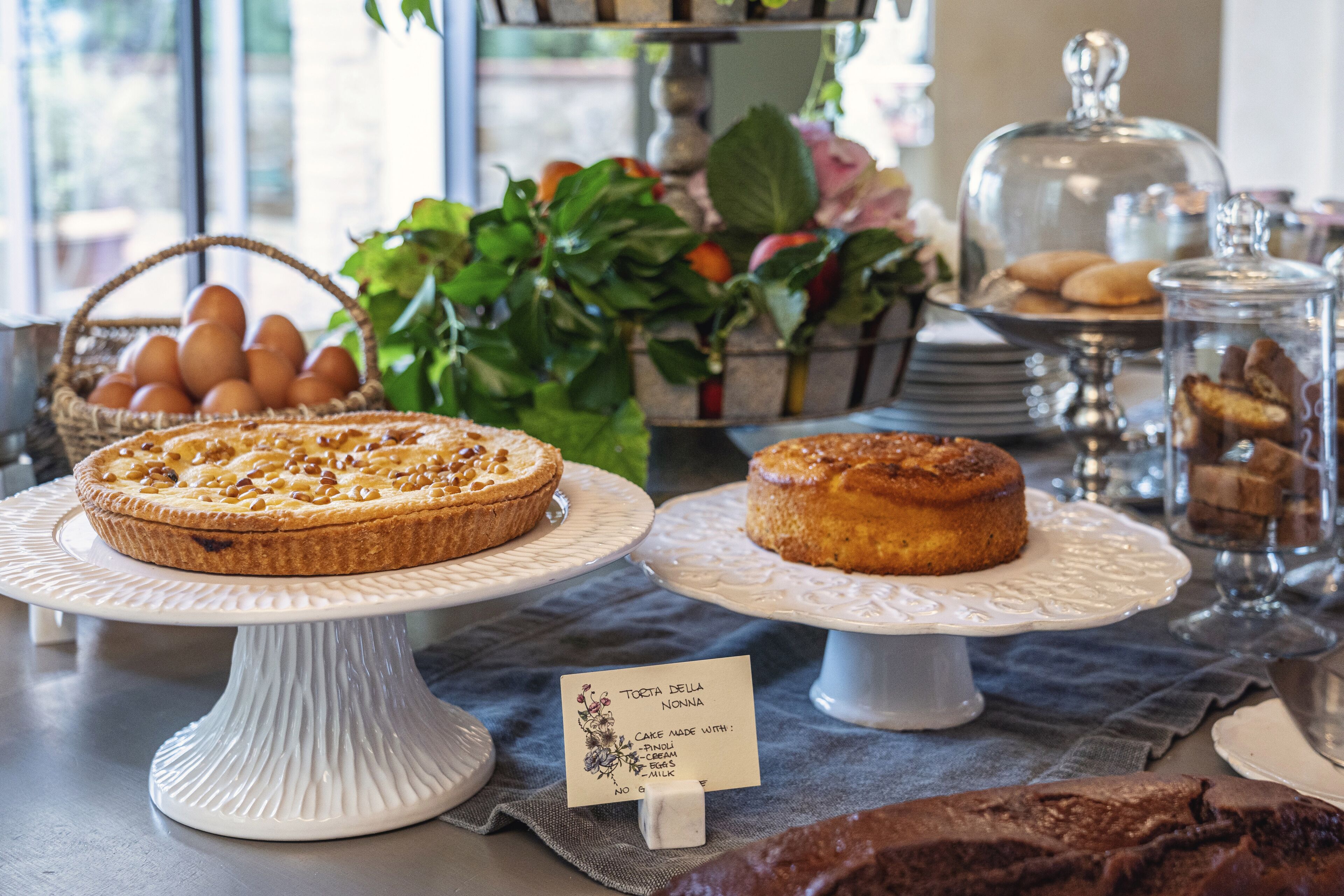 Una vitrina de pastelería con varios pasteles y productos de repostería sobre bases blancas, una cesta de huevos y un letrero que indica un pastel hecho con leche.