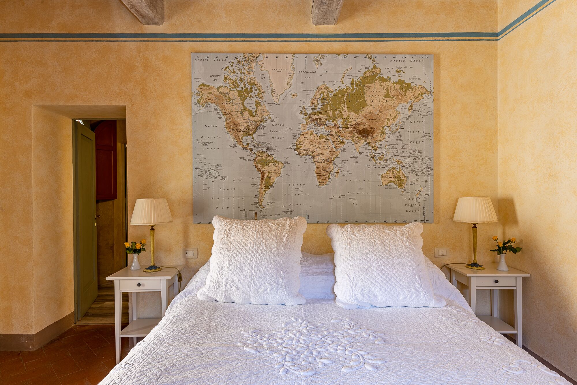 A bedroom with a large world map on the wall, a bed with white linens, two bedside tables with lamps, and a door leading to another room.