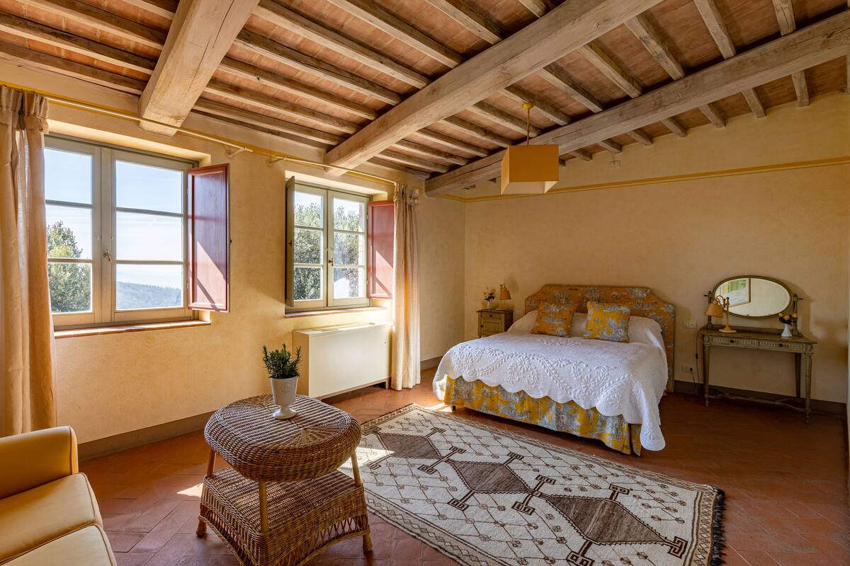 A bedroom with a bed, a small table with a plant, a wicker chair, and a patterned rug.