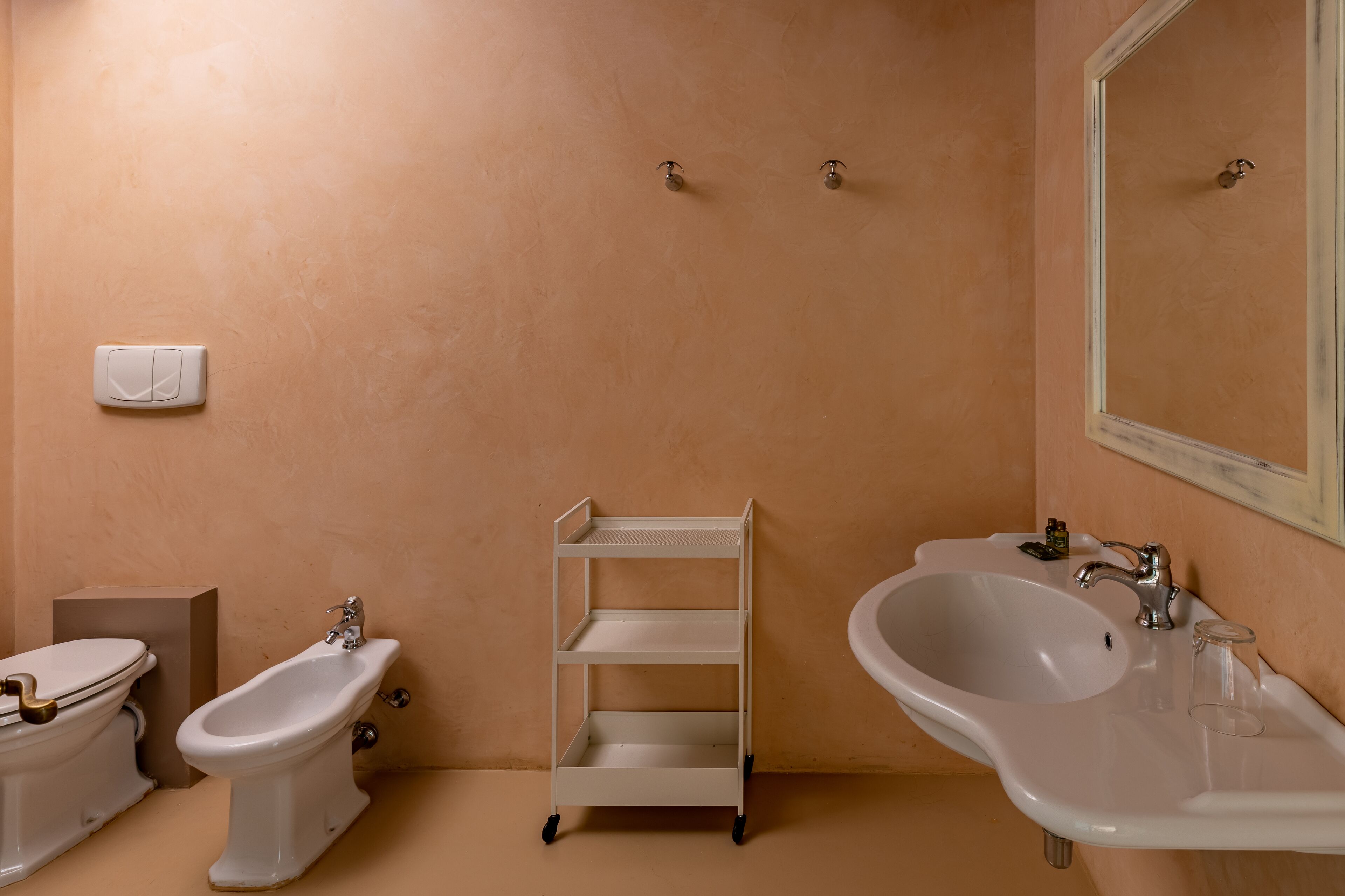 A bathroom with a toilet, bidet, shelf, sink, and mirror.