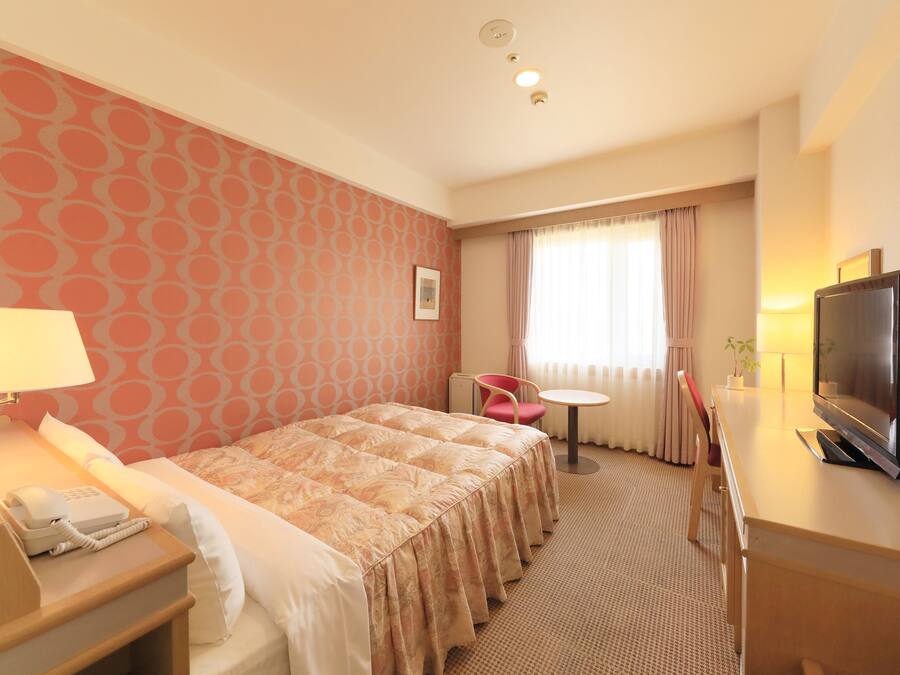 A hotel room with a bed, bedside table with a lamp and phone, a TV, a small table with a chair, and a window with curtains.