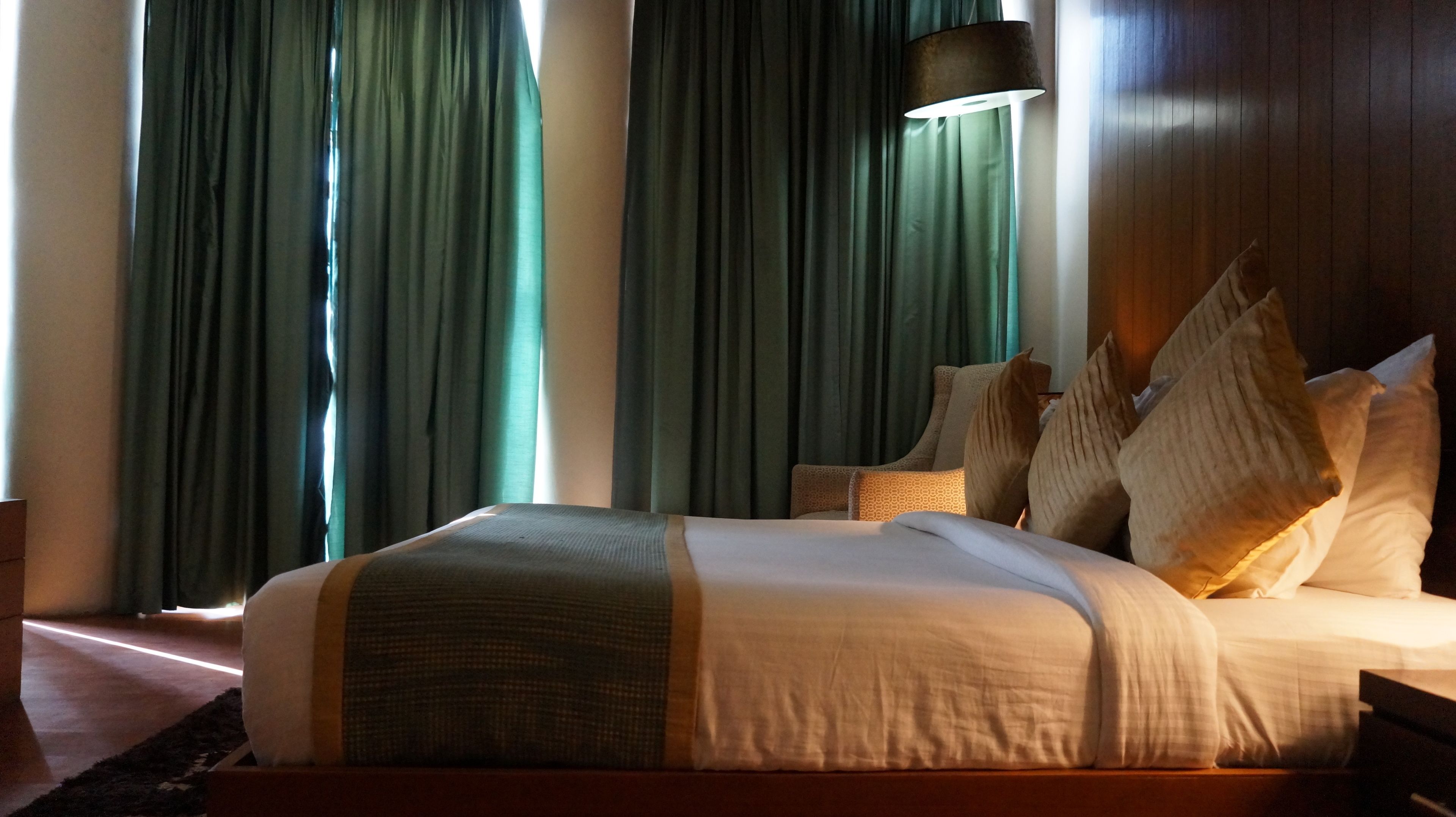 A hotel room with a large bed, green curtains, a standing lamp, and a wooden headboard.