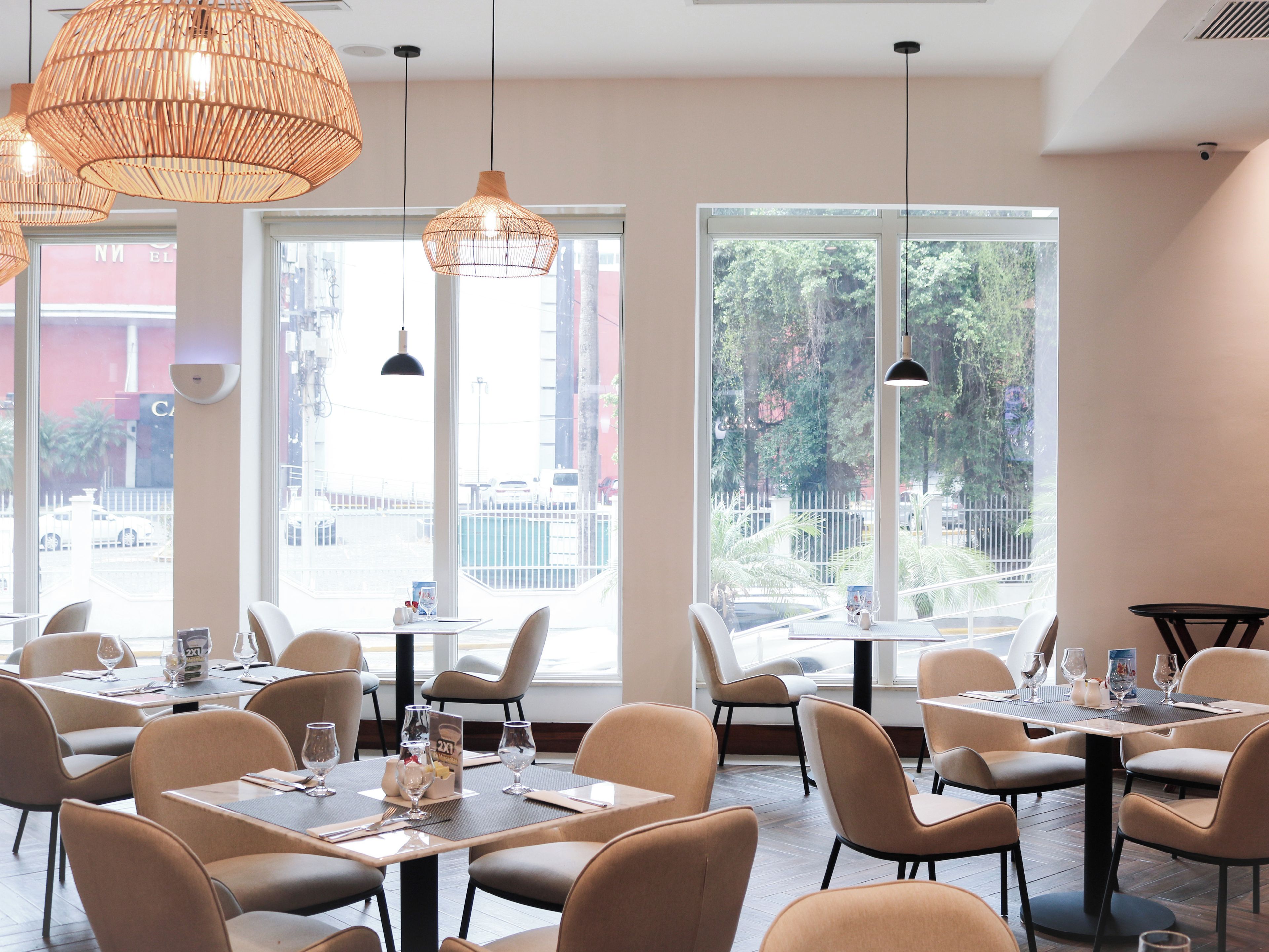Interior de un restaurante con varias mesas y sillas, amplias ventanas con vistas al exterior e iluminación colgante.
