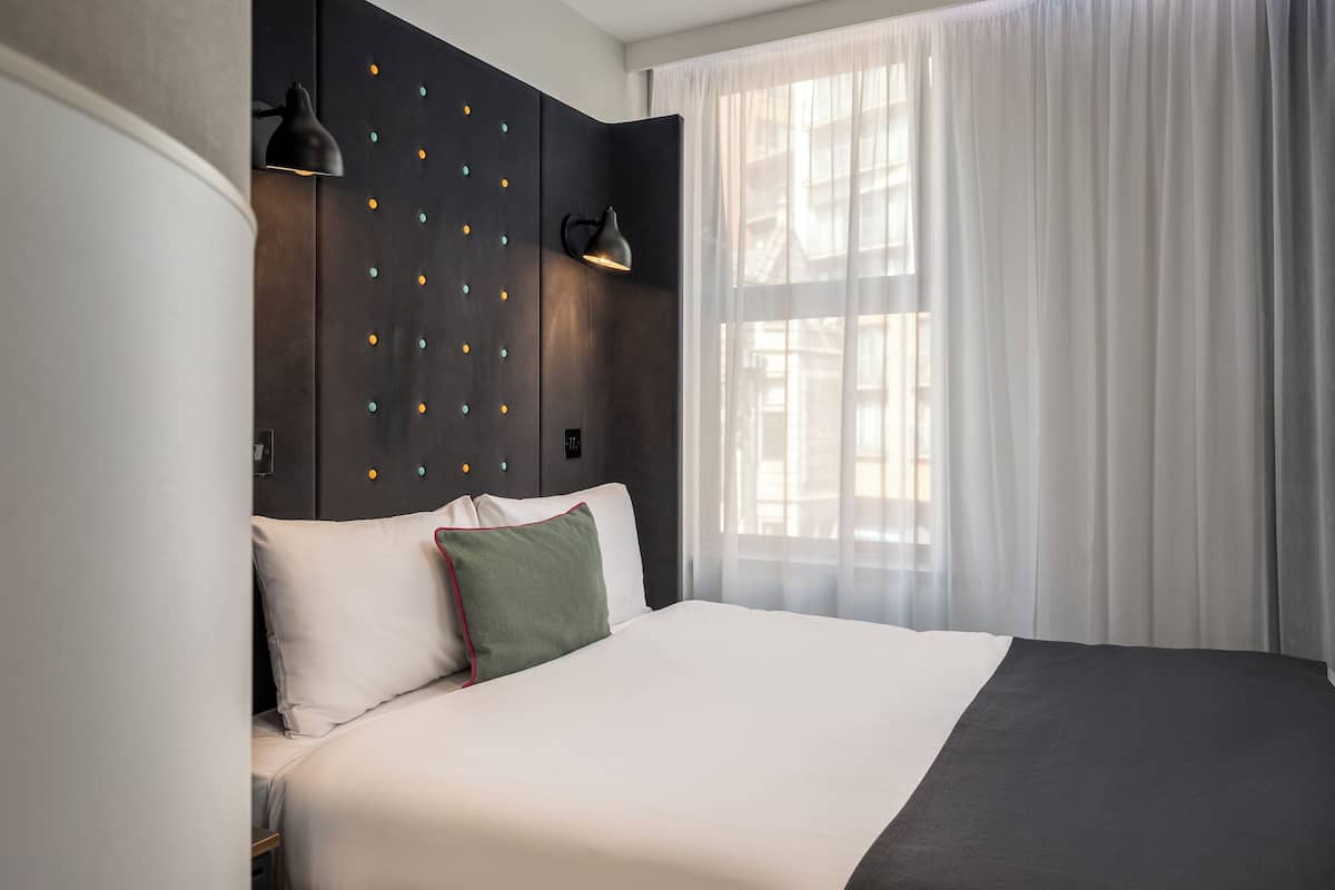Una habitación de hotel moderna con una cama grande, un cabecero oscuro con luces decorativas, una lámpara de noche y cortinas traslúcidas.