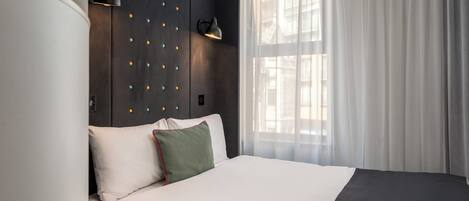 Una habitación de hotel moderna con una cama grande, un cabecero oscuro con luces decorativas, una lámpara de noche y cortinas traslúcidas.